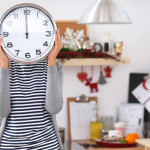 A woman standing in front of a kitchen area, holding a clock in front of her face