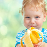 A smiling young child looking directly into the camera, holding a partially eaten banana towards the viewer