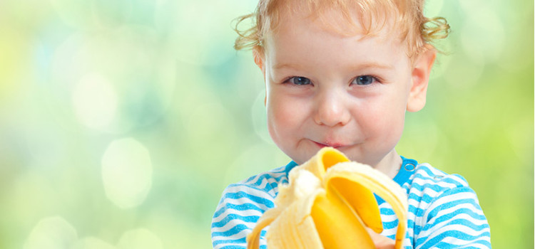 A smiling young child looking directly into the camera, holding a partially eaten banana towards the viewer