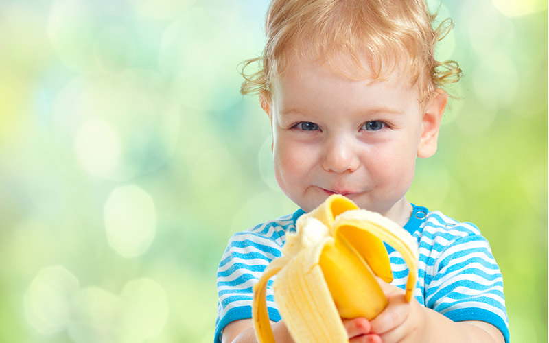 A smiling young child looking directly into the camera, holding a partially eaten banana towards the viewer