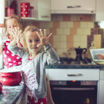 A home cooking kitchen scene with an apron wearing mother in the background and 2 young kids helping out in the foreground. Holding up dough covered hands to the viewer. Kitchen is a bit of a mess.