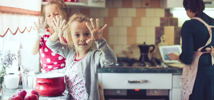 A home cooking kitchen scene with an apron wearing mother in the background and 2 young kids helping out in the foreground. Holding up dough covered hands to the viewer. Kitchen is a bit of a mess.