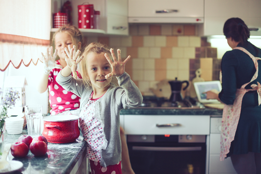 A home cooking kitchen scene with an apron wearing mother in the background and 2 young kids helping out in the foreground. Holding up dough covered hands to the viewer. Kitchen is a bit of a mess.