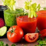 Four glasses with vegetable juices in them sitting on a table surrounded by a beet, and cucumber a tomato and a carrot.