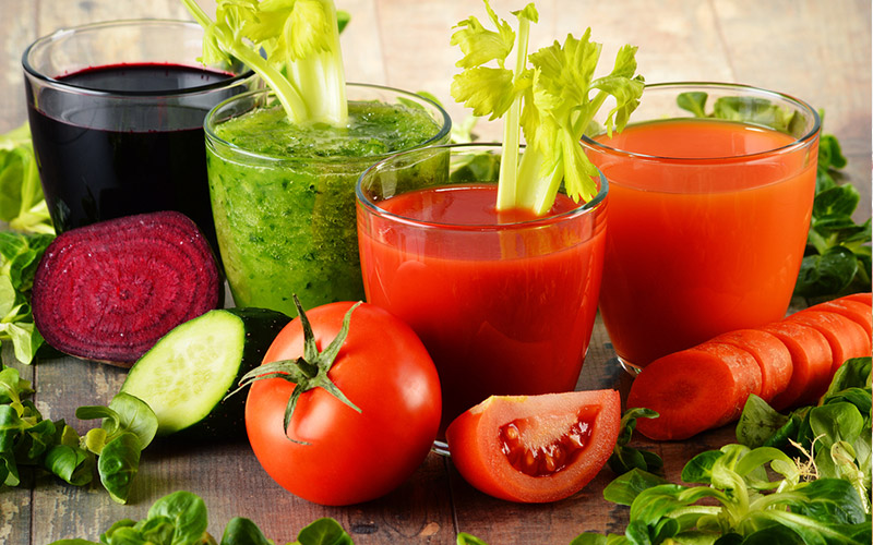 Four glasses with vegetable juices in them sitting on a table surrounded by a beet, and cucumber a tomato and a carrot.