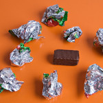 An orange surface with 9 crumpled silver candy wrappers and a single chocolate candy