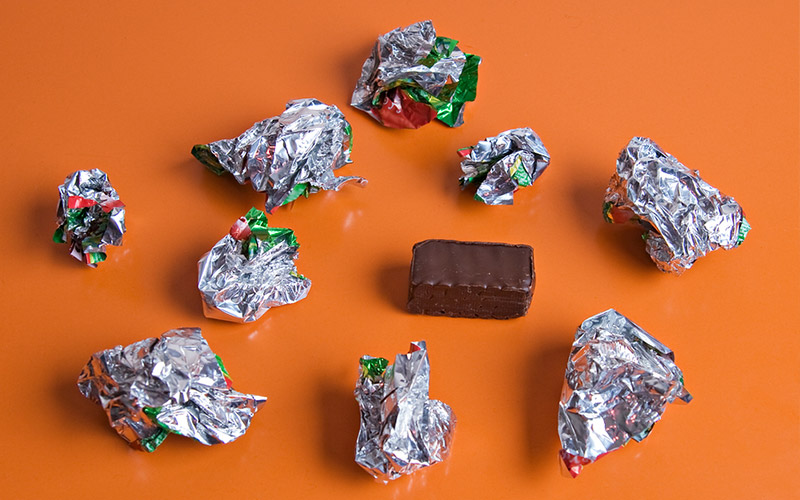 An orange surface with 9 crumpled silver candy wrappers and a single chocolate candy