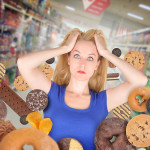 A harried looking woman in a blue t-shirt, grabbing clumps of her hair and staring at the viewer- surrounded by all forms of junk food that is flying around her.