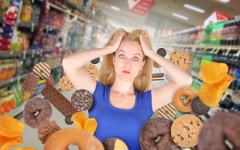 A harried looking woman in a blue t-shirt, grabbing clumps of her hair and staring at the viewer- surrounded by all forms of junk food that is flying around her.