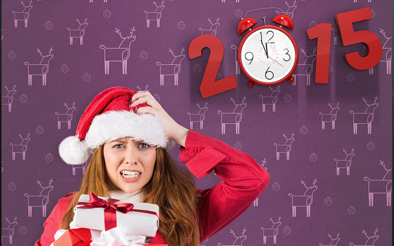 A woman in a santa outfit scratching her head while holding an armful of wrapped gifts. An old fashioned alarm clock is in the background forming the '0' part of a red numbered "2015"