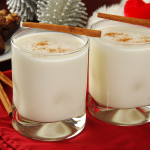 2 glass cups of eggnog with cinnamon sticks on the side, on a festive Christmas themed table