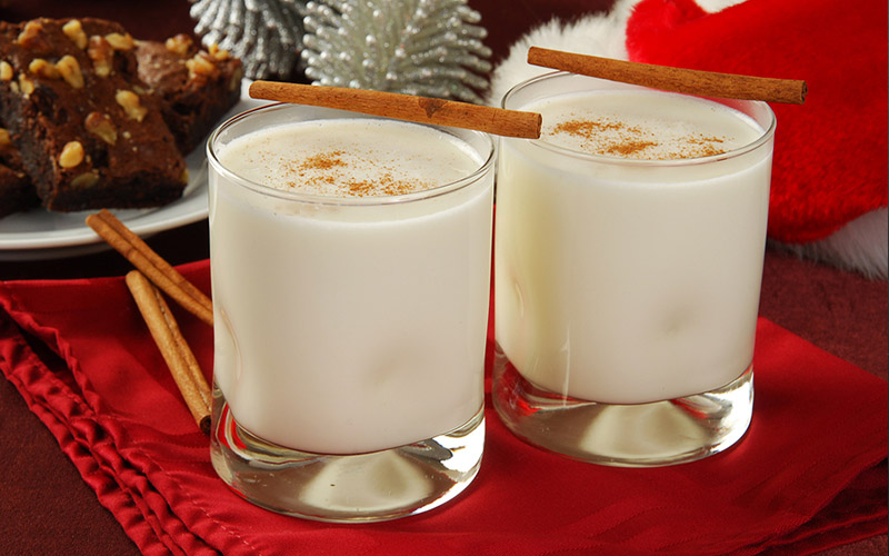 2 glass cups of eggnog with cinnamon sticks on the side, on a festive Christmas themed table