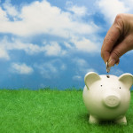 White ceramic piggy bank with a woman's hand dropping a coin into it. All on a green grass ground with partially cloudy sky in the background