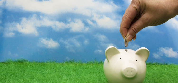 White ceramic piggy bank with a woman's hand dropping a coin into it. All on a green grass ground with partially cloudy sky in the background