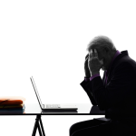 An older man leaning on his hands which are pressed against his temple. He is having a hangover in front of his laptop at work.