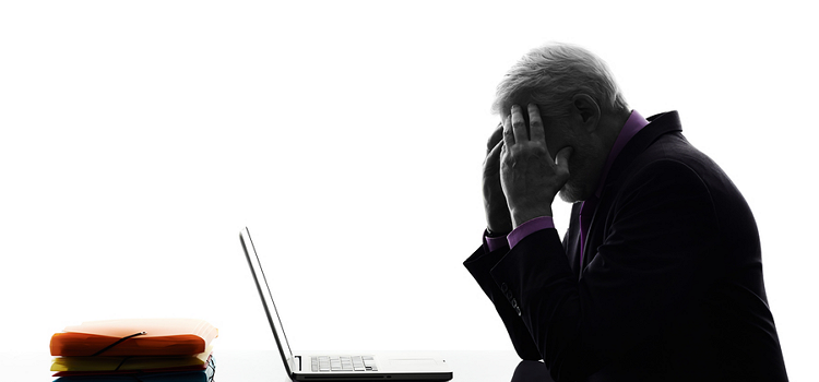 An older man leaning on his hands which are pressed against his temple. He is having a hangover in front of his laptop at work.