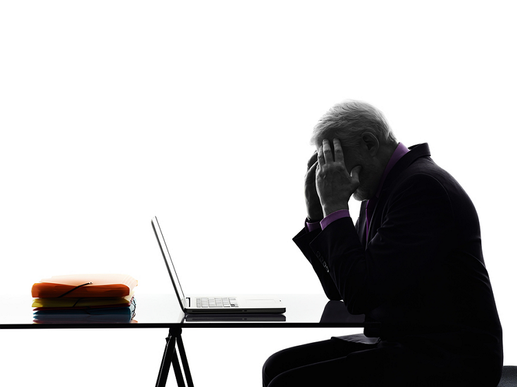 An older man leaning on his hands which are pressed against his temple. He is having a hangover in front of his laptop at work.